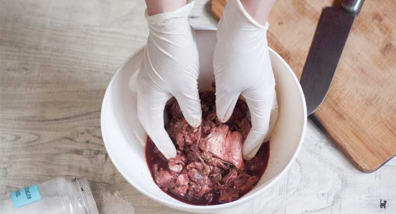 Hände in Einmalhandschuhen mischen Rohfleischmahlzeiten in einer kleinen weißen Plastikschüssel